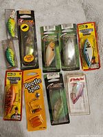 Top view of assorted new fishing lures in original packages laid flat on white textured surface showing brand names and lure colors