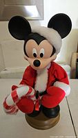 Front view of Mickey Mouse dressed as Santa holding a large candy cane, on a brown base.