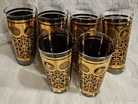 Group of six tall cylindrical glasses featuring black and gold decorative panels with scrollwork and medallion patterns.