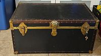Front view of rectangular black storage trunk showing brass lock, clasps, rivets, and key attached to lock.
