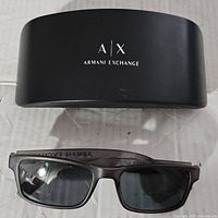Black hard sunglasses case with Armani Exchange logo and matching Armani Exchange black sunglasses with rectangular lenses shown on white background.