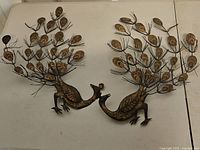 Pair of vintage metal peacock wallhangings shown resting on table, front view.