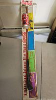 Full view of the Coca-Cola fishing rod in original packaging, showing colorful bottle illustrations along the package length and the fishing rod extending from top to bottom.