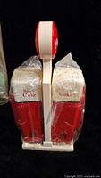 Front view of two Coca-Cola salt and pepper shakers wrapped in plastic, placed in white plastic caddy holder with Coca-Cola logo on top.
