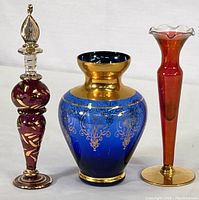 Image of three glass items: ruby red perfume bottle with stopper, blue and gold vase, cranberry fluted bud vase on a white background.