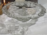 Footed glass cake plate with raised floral decorations and scalloped rim alongside two etched glass dessert cups on a white surface.