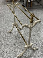 Full view of vintage Victorian style quilt rack with cast enameled metal frame and brass bars standing on gray speckled floor.