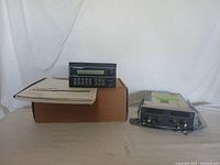 Two aviation electronic devices positioned side by side on a table, one black rectangular unit with buttons and screen, the other a silver metal box with control knobs and connectors, also shows a cardboard box under the black unit.