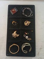 Full view of assorted used jewelry in a black display box divided into multiple compartments showing brooches, bracelets, earrings, and necklace.