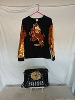 Full lot showing black ASU PINK blouse with gold sequin sleeves hanging on pink hanger above black Steve Madden handbag with gold sequin lips and 'FABULOUS' on front flap.