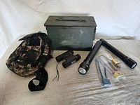 Full lot shot displaying camouflage backpack, large green metal ammo box, two black flashlights, black binoculars, multiple packs of tent stakes and matches on white surface.