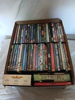 Top view of two rows of assorted DVD movies in original cases inside a cardboard box showing colorful spines and various titles.