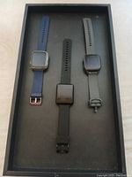 Three fitness watches in tray, one with blue band and rose gold buckle, one black, one grey.
