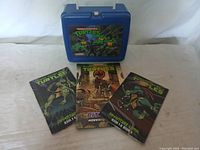 Blue Teenage Mutant Ninja Turtles lunch box with three magazines laid out in front showing different comic or adventure-themed covers with TMNT characters.