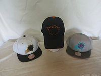 Three Phoenix Suns hats positioned side by side showcasing different colors and logos: white/black, black/orange, and gray/teal-pink.