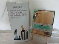 Photo of the Secura stainless steel electric wine opener and ice bucket gift set in original packaging alongside the wooden cheese board with spreader set in packaging on a small wooden stand.