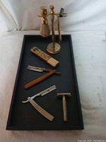 Overview of the grooming tools on a black rectangular tray including shaving brush with gold-tone stand, two straight razors, safety razor and accessory case.