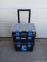 Three stacked black plastic storage containers with blue latches and clear plastic lids, with a black retractable handle, positioned outdoors against a garage door.