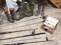 Photo showing three pairs of fishing boots: tall green waders, Original Muck Co boots, and grey Grundens boots; five fishing rods with reels and a tackle box containing fishing accessories