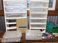 Two white plastic drawer units on a table near window, one with missing drawers, small clear plastic storage boxes in front, red velvet sheet behind, and some craft items to side.