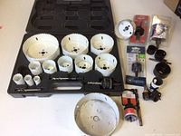 Set of white-coated hole saws arranged inside a black plastic case with various sizes, plus additional loose hole saws and accessories outside the case.
