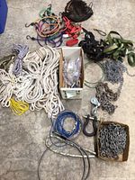 Wide overview photo showing multiple ropes, chains, ratchet straps, bungee cords, steel cables, and a cardboard box containing chains and a pulley.