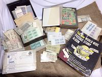 Multiple envelopes containing postage stamps, with partial view of two albums. One album is large and black with text, the other blue. Various stamps visible in envelopes and pages.