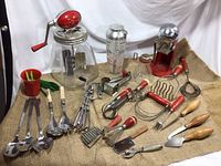Overview of the entire lot showing red vintage kitchen appliances including an ice crusher, a hand mixer, and various hand utensils with wooden and red handles arranged on a burlap cloth.