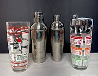 Photo showing two stainless steel cocktail shakers and two tall cocktail glasses with printed retro cocktail recipes and illustrations, set against a dark background.