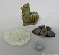 Four hand carved jade pieces displayed on a light surface: brown-green seated animal, translucent double koi fish, dark flower-shaped pendant, small light green Buddha pendant.