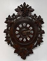 Front view of the antique carved oak German wall clock showing carved leaves and grape design surrounding the clock face with white Roman numeral markers and decorative clock hands.