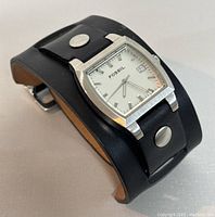Side angle showing Fossil watch with square silver stainless steel case and black leather cuff band with metal rivets.