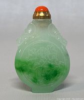 Front view of the hand carved jade perfume/snuff bottle showing its gradient green and white color with detailed carving and coral cap.