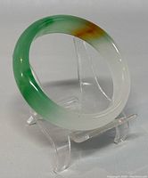 Photo of the natural jade bangle on clear stand showing green, white, and brown colors and D-shape profile.