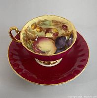 Angle view showing the teacup with hand-painted fruit inside and red exterior with gold trim, plus matching saucer of a different red shade