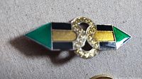 Close up top-down image of an Art Deco style brooch with green, black, and gold-tone enamel shapes and a central gold ornate section, laid on a beige fabric background.