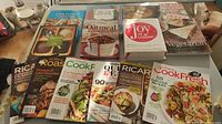 Photo of cookbooks and cooking magazines arranged on a surface, including titles: Spanish, Oatmeal Cookbook, Joy of Cooking, Vegetarian, and various Canadian cooking magazines.