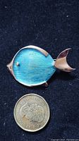 Front view of fish brooch with turquoise coloured body and gold tone metal edges, next to a coin for size reference.