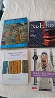 Four needlecraft books arranged on white bedding showing titles and cover images of Glorious Needlepoint, Sashiko, Japanese Stitches Unraveled, and Japanese Knitting.