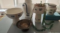 Front and side angle view of ceramic vases and bowls with abstract face designs and trailing succulent.
