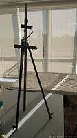 Front view of black metal easel showing tripod legs, vertical pole, and horizontal clamp for canvas support.