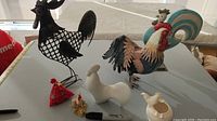 View showing seven rooster and chicken themed items displayed on a white table, including metal and ceramic figurines and a fabric pincushion.
