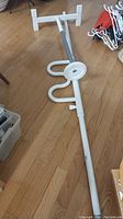White metal security pole with curved hook section, flat foot plate at the base, and attached visegrip clamp laying on wood floor.