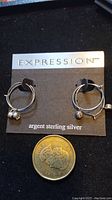 Two pairs of earrings on the Expression branded card, next to a coin for size reference. Both pairs are sterling silver hoops with different charms.