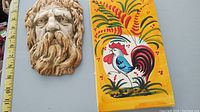 Photo showing bearded face wall hanging with a measuring tape beside it and ceramic tile with rooster design