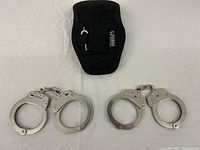 Photo showing two metal handcuff sets side by side with a black nylon pouch above them on a white background.