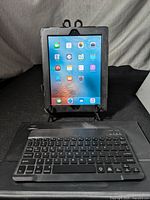 Image of Apple iPad 2 in black protective case showing home screen with apps and black wireless keyboard in front of it.
