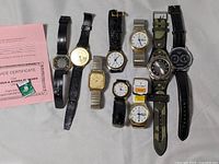 Full lot of 9 assorted wristwatches arranged in a row showing dials and straps, including a pink service certificate and battery on side.