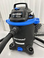Front angle showing Mastervac shop vacuum with hose attached, blue secure latch, and caster wheels.