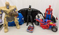 Thanos, Batman, Spiderman action figures, Spiderman motorcycle vehicle toy, two blankets in background, Spiderman mini puzzle tin on table.